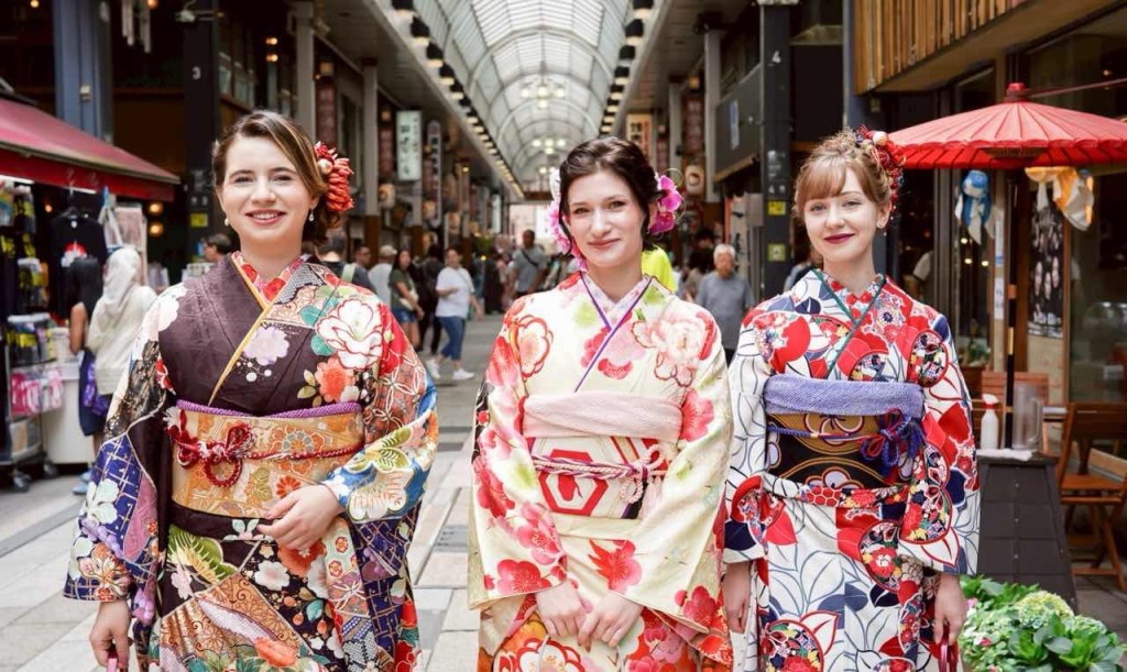 日本の伝統的な着物を着た3人の女性が、賑やかな商店街を背景に笑顔で並んでいる様子