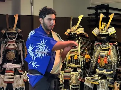 A performer in traditional Japanese attire gestures while standing in front of samurai armor displays.