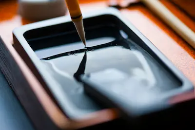 A calligraphy brush being dipped into a black ink tray, with soft reflections on the ink surface.