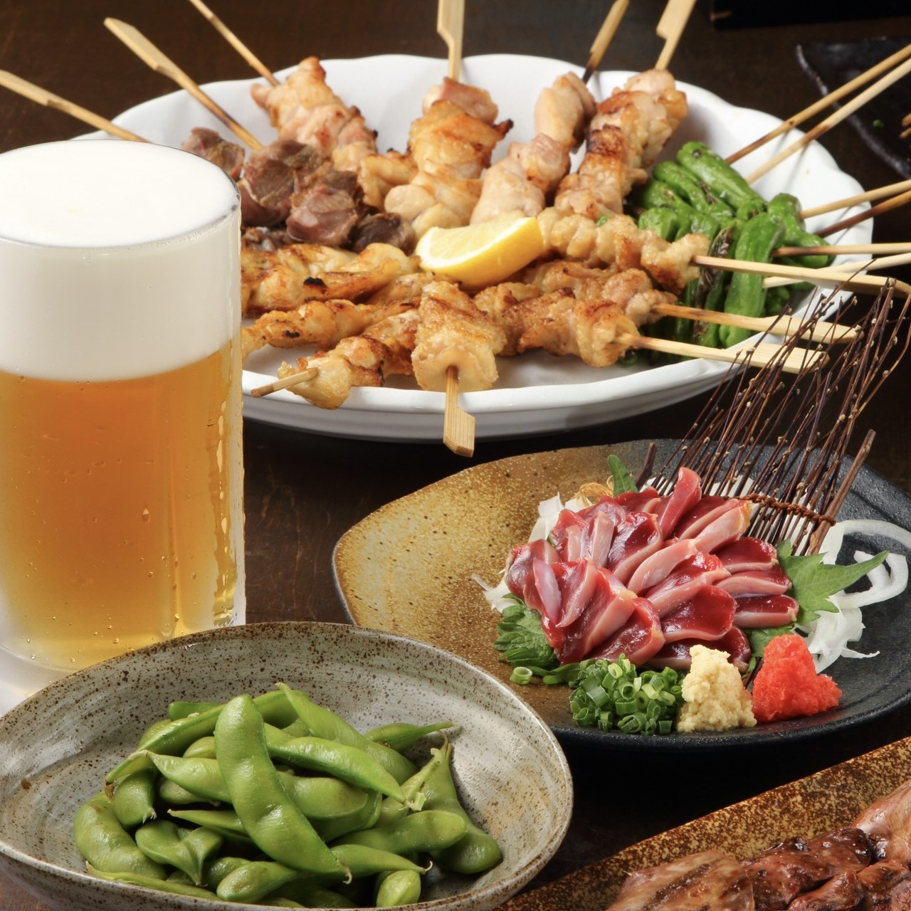 A selection of Japanese izakaya dishes including grilled skewers of meat, served with edamame, sashimi, and a cold beer.