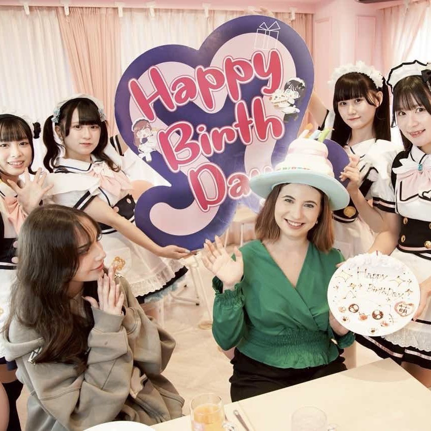 A joyful birthday celebration at a themed café in Japan, featuring a woman with a green top holding a birthday cake and several waitresses in costume, alongside a festive 'Happy Birthday' sign.