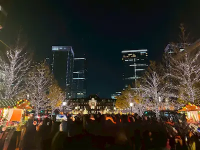 A bustling winter scene in Tokyo at night, filled with illuminated trees and vibrant lights, showcasing tall buildings in the background and a crowd enjoying the festive atmosphere.