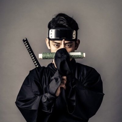 A person dressed in traditional ninja attire, holding a rolled scroll and standing against a neutral background.
