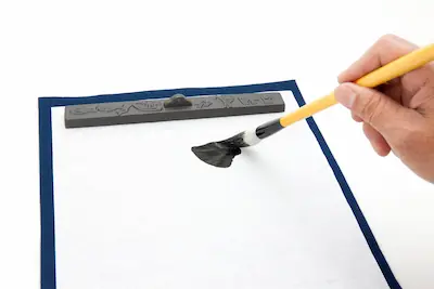 A hand holding a brush is applying black ink to a blank sheet of paper, with an inkstone visible on the edge.