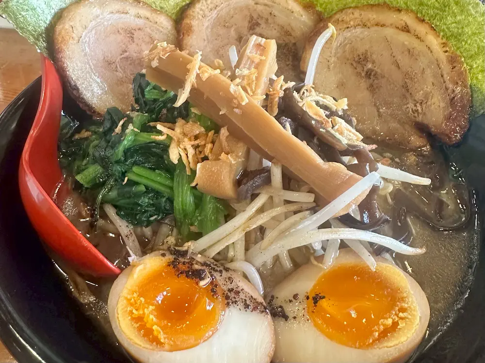 A bowl of ramen featuring thick slices of pork, soft boiled eggs with runny yolk, fresh greens, and bamboo shoots, presented with a vibrant red spoon.