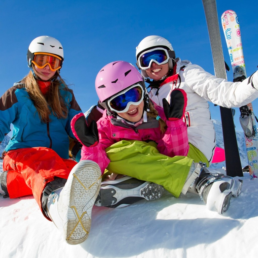 Three children enjoying a day in the snow, wearing colorful ski outfits and goggles, posing happily while sitting on a snowy slope under a clear blue sky.