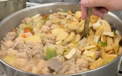 A hand stirring a large pot of chanko nabe, a traditional sumo wrestler hotpot, filled with meat and vegetables.
