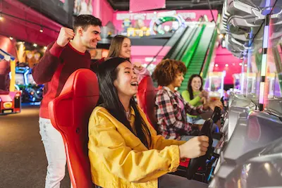 Group of friends enjoying a racing arcade game in a colorful arcade setting. One person is excitedly driving while others cheer them on.