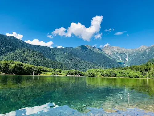 Serene view of a clear lake surrounded by lush green mountains under a bright blue sky with fluffy clouds.