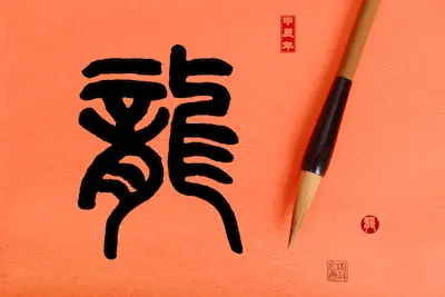 Traditional Japanese calligraphy featuring an intricate kanji character on an orange background, accompanied by a brush.
