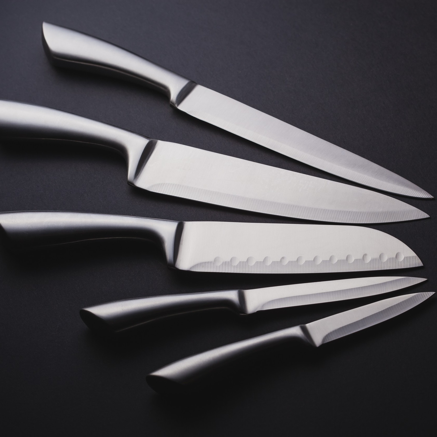 A collection of five stainless steel kitchen knives arranged on a dark surface, showcasing various blade shapes and sizes.