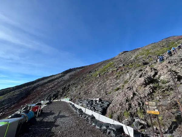A steep hiking trail leading up a rocky slope, lined with safety barriers and surrounded by vibrant greenery under a clear blue sky.