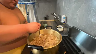 A sumo wrestler stirring a large pot of chanko nabe in a kitchen setting.