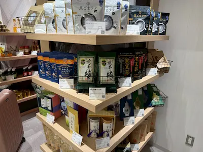 A wooden display shelf stocked with various Japanese products, including snacks, beverages, and traditional goods, with clear labels and price tags.