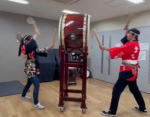 Two performers engaging in a taiko drumming session, one wearing a traditional Japanese outfit and the other in casual attire, both striking a large taiko drum with drumsticks in a practice room.