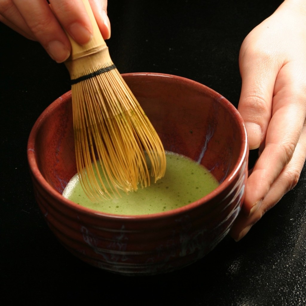 A close-up of a hand whisking matcha tea with a bamboo whisk in a traditional ceramic bowl.
