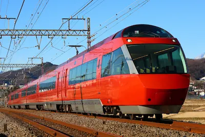 A red Odakyu Romancecar train traveling on tracks with overhead electrical lines, surrounded by mountainous scenery.