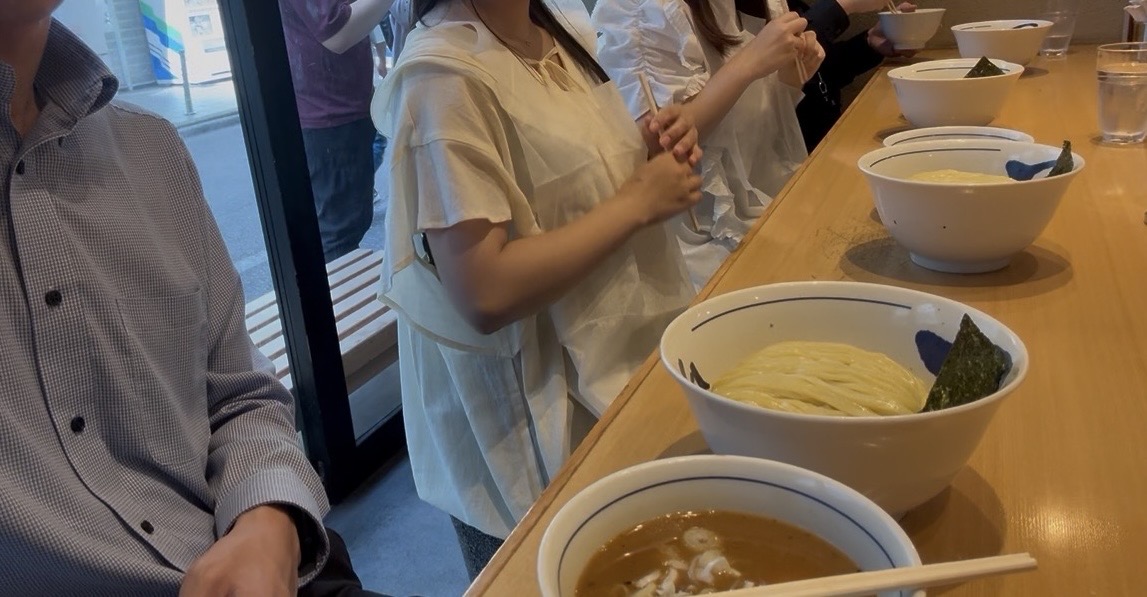 Customers sitting at a wooden counter in a ramen shop, wearing plastic aprons, with bowls of ramen in front of them.