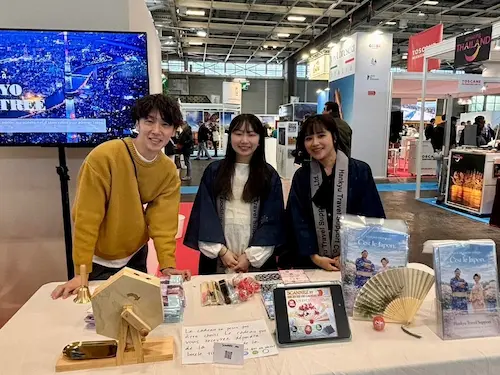 展示会で笑顔の3人がブースで写真を撮っている。テーブルには観光案内やパンフレット、扇子が並べられている。